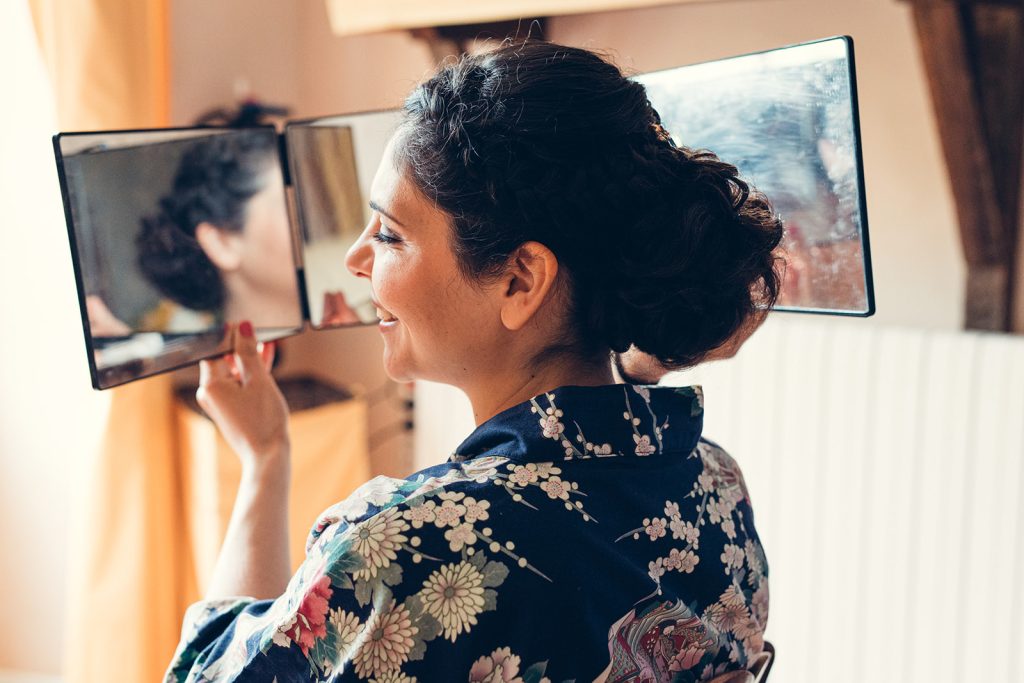 La mariée est heureuse de son maquillage et de sa coiffure en se voyant dans le miroir lors des préparatifs de son mariage au relais de porthos à Ogenne-Camptort
