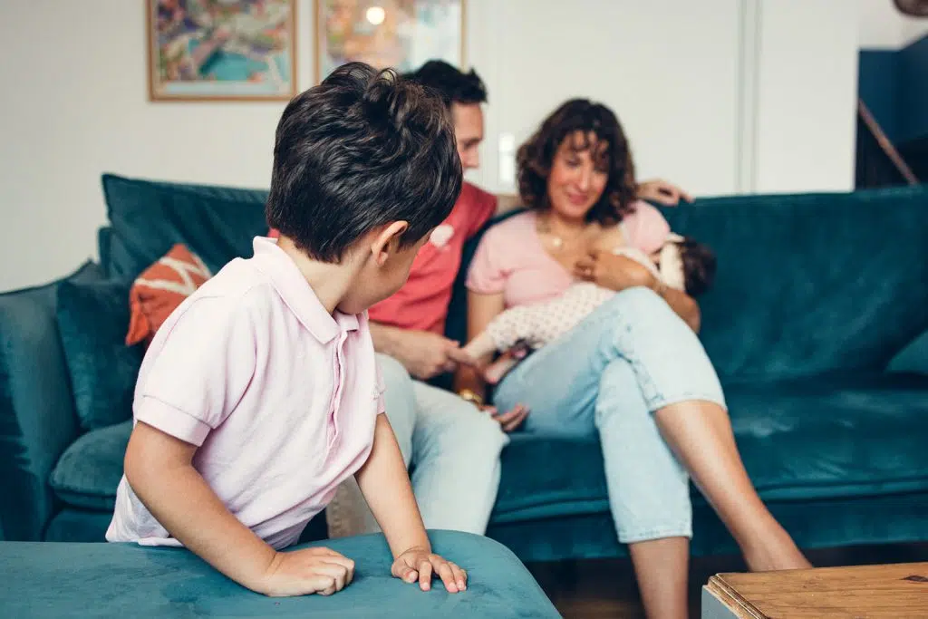 le grand frère regarde ses parents assis sur le canapé du salon bercer sa petite soeur lors d'une séance naissance à domicile à nay