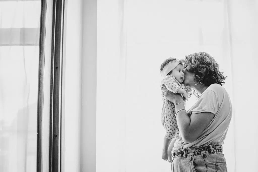 la maman embrasse tendrement son bébé devant la fenêtre de son salon lors d'une séance naissance à domicile à nay